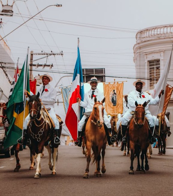 Três Mantenas a cavalo, de terno e chapéu brancos, levam a bandeira do Brasil, a de Guaratinguetá e estandartes pela rua