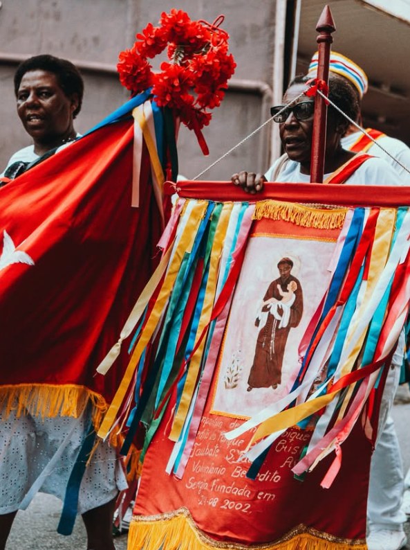 Devotas carregam um estandarte vermelho com a imagem de um santo frade com o Menino Jesus, enfeitado com fitas coloridas e cravos vermelhos