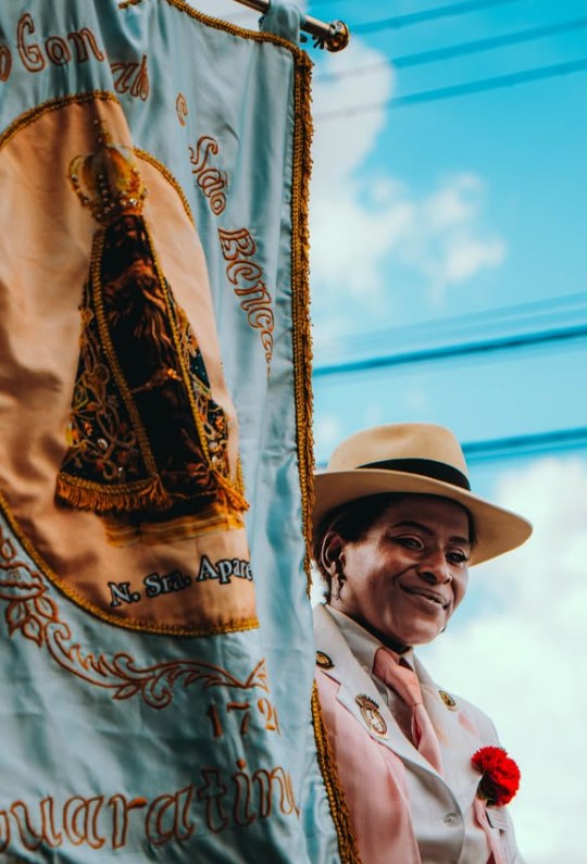 Uma Mantena de chapéu, lenço rosa e cravo vermelho sorri ao lado do estandarte de Nossa Senhora Aparecida